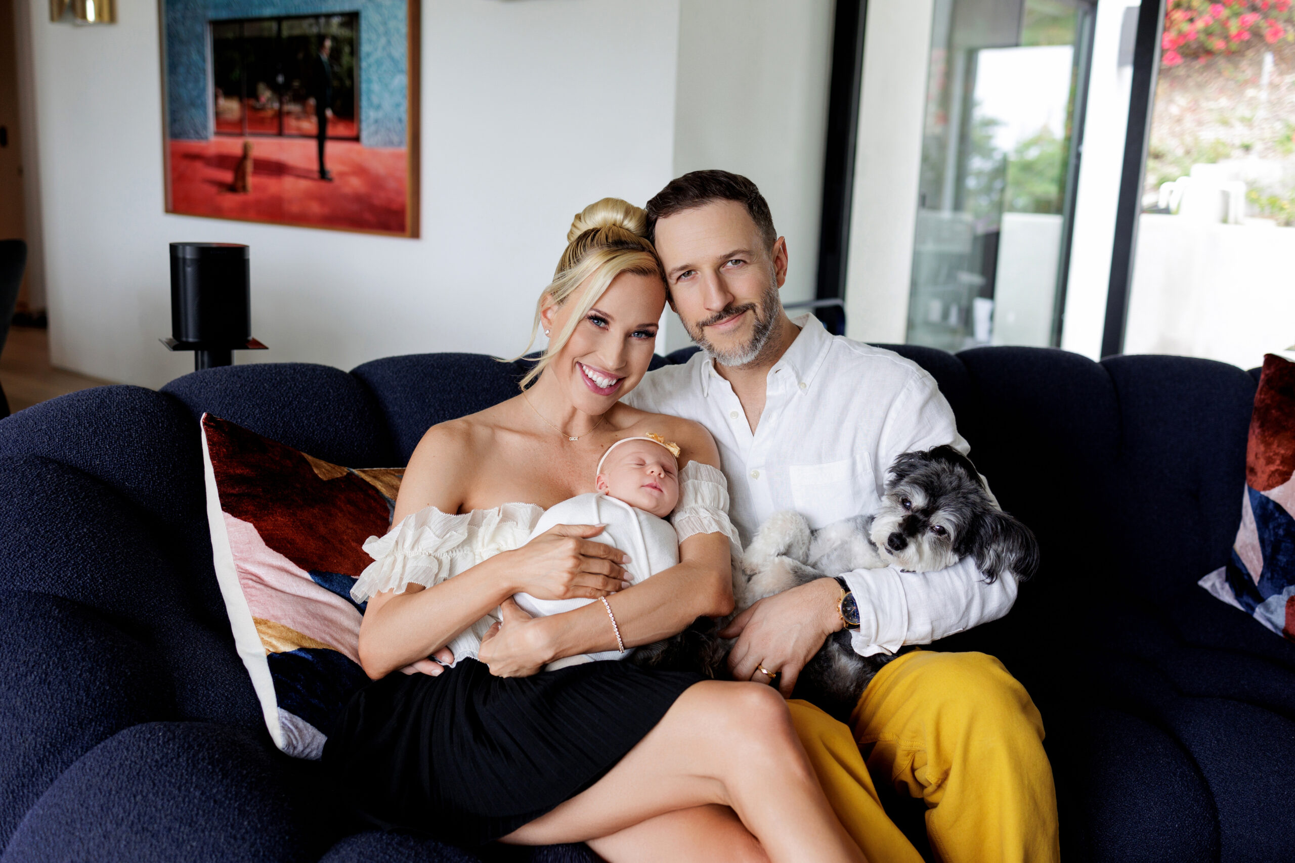 A happy mother and father hold their baby and cuddle their dog while sitting on their couch.