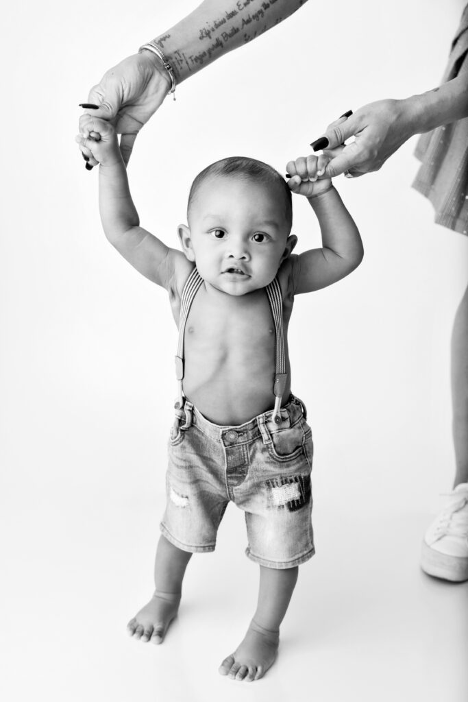 Baby wearing only overalls walking assisted by mom on a white backdrop at a North Hollywood Photography studio