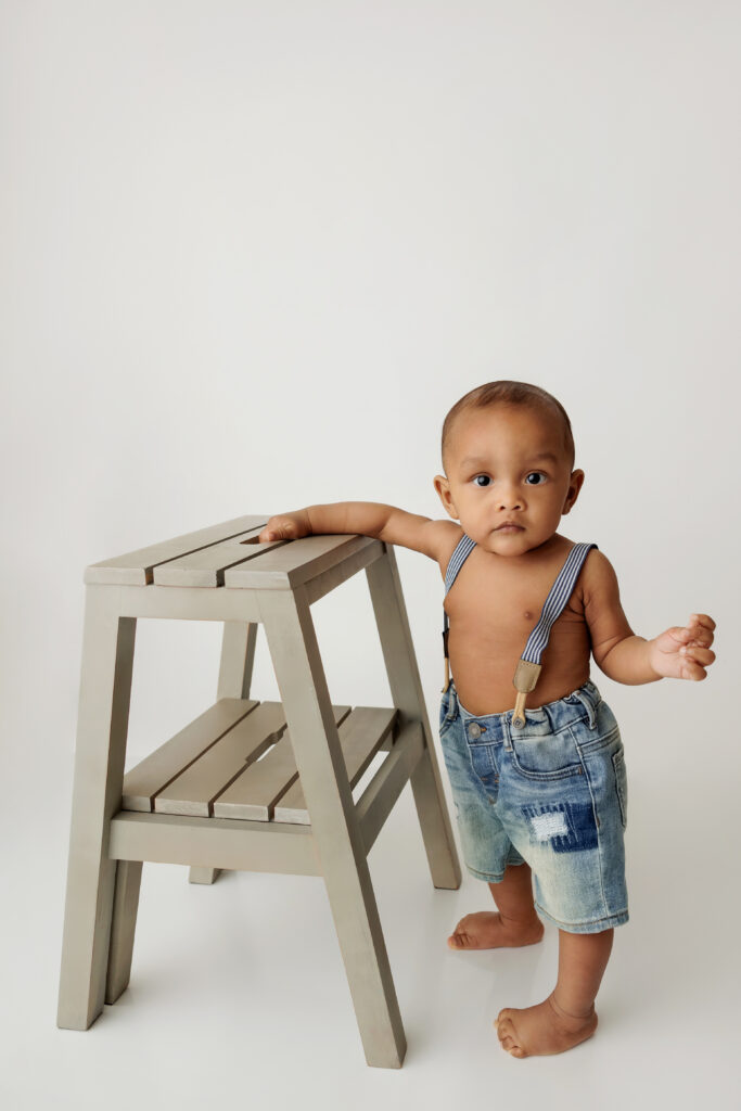 Baby wearing only overalls holding onto a small step ladder on a white backdrop at a North Hollywood Photography studio